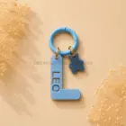 Chapa identificativa personalizada para perros con inicial L azul cielo, nombre Leo troquelado y colgante decorativo de osito sobre fondo beige.