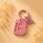 Chapa identificativa personalizada con inicial para perros en color rosa, nombre troquelado BELLA, colgante mariposa y mosquetón rosa sobre fondo beige decorativo.