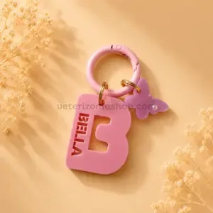Chapa identificativa personalizada con inicial para perros en color rosa, nombre troquelado BELLA, colgante mariposa y mosquetón rosa sobre fondo beige decorativo.