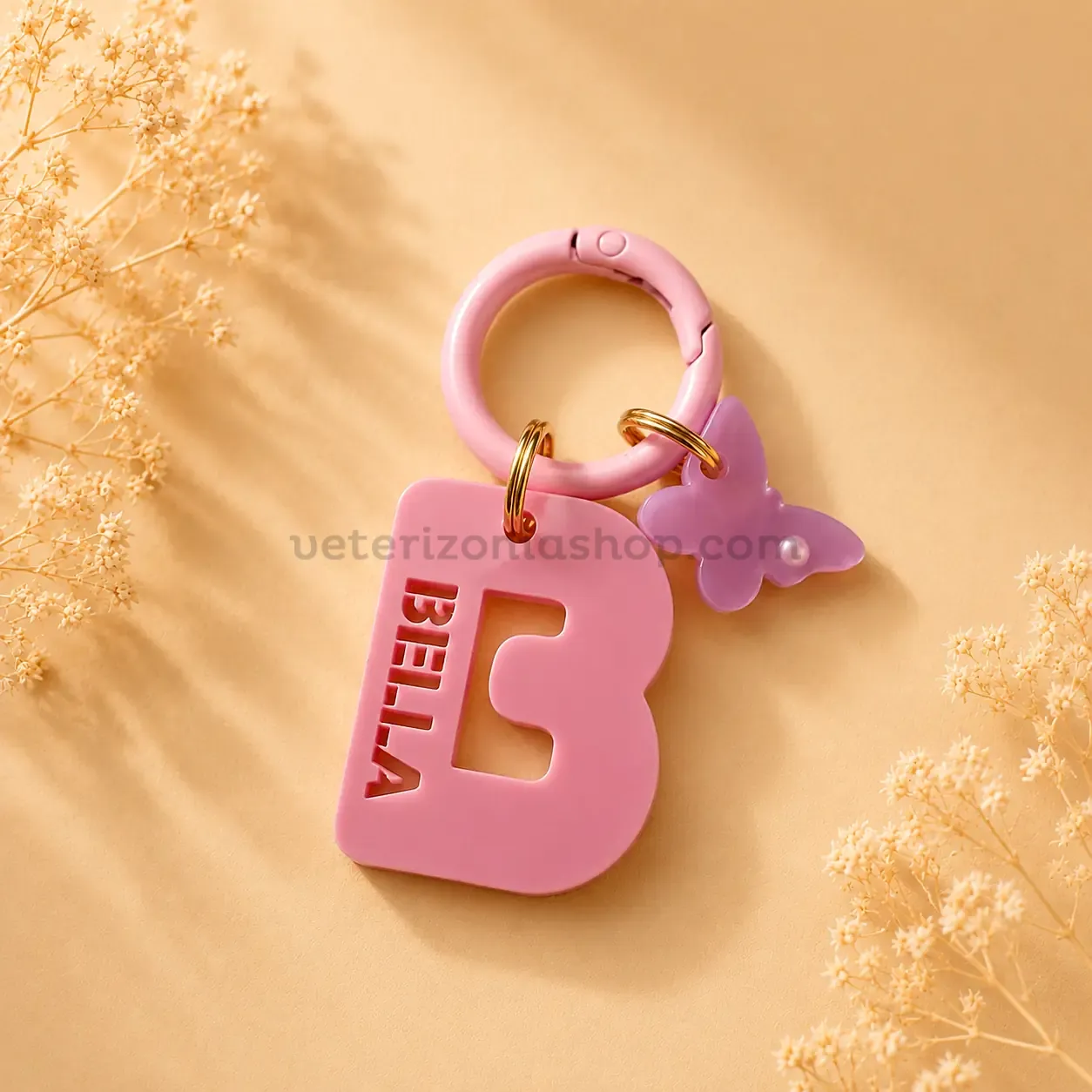 chapa-identificativa-personalizada-inicial-perros-bella Chapa identificativa personalizada con inicial para perros en color rosa, nombre troquelado BELLA, colgante mariposa y mosquetón rosa sobre fondo beige decorativo.